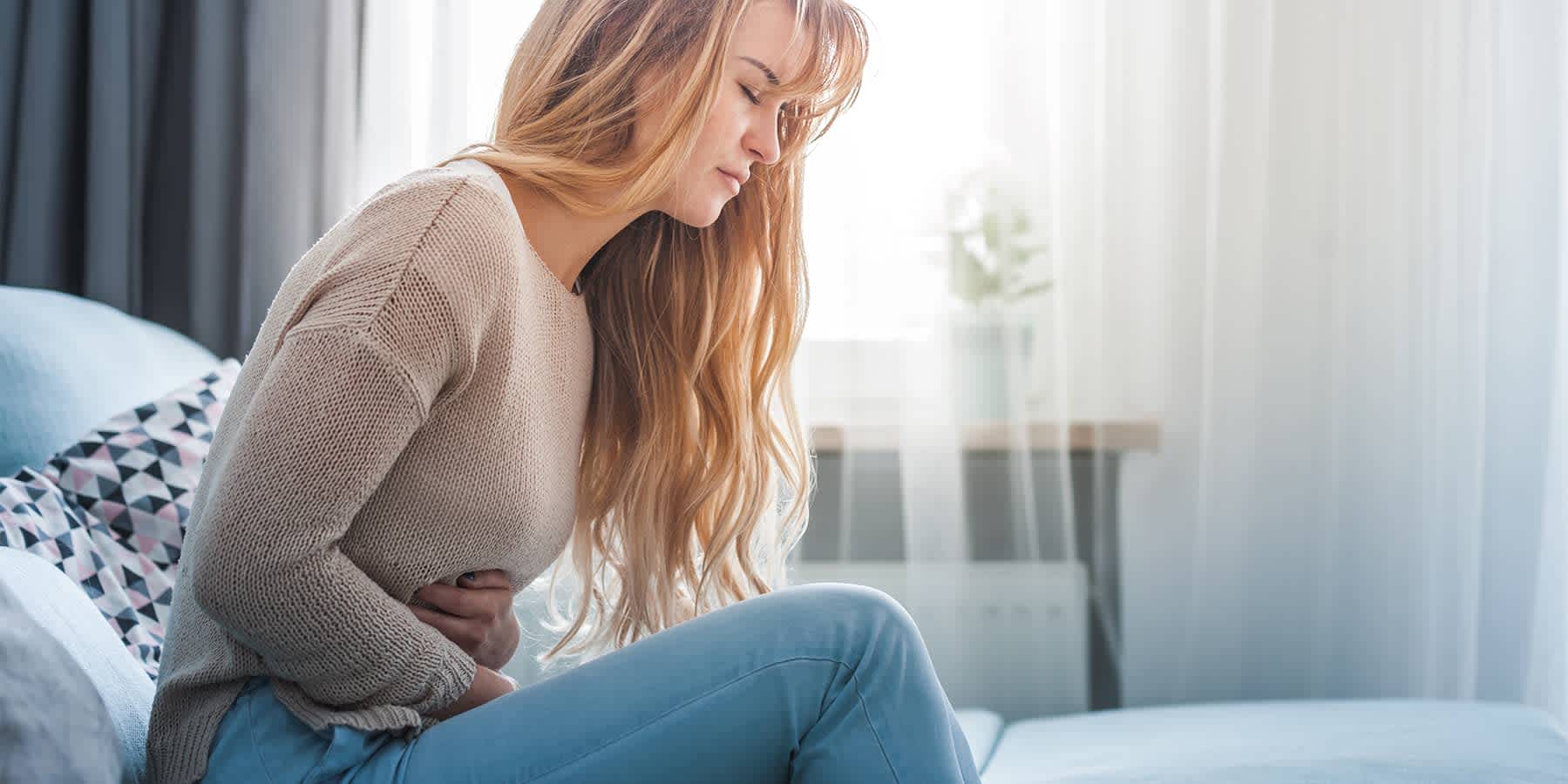 Woman sitting on bed with hands on her stomach while experiencing an IBS flare up